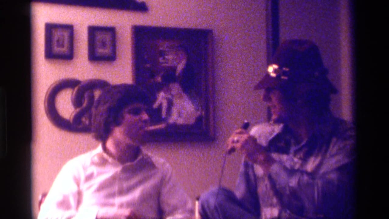 In a 1976 Kentucky 8mm home movie, two adult men sit in a dim room as the man in a hat holds a microphone toward his friend.