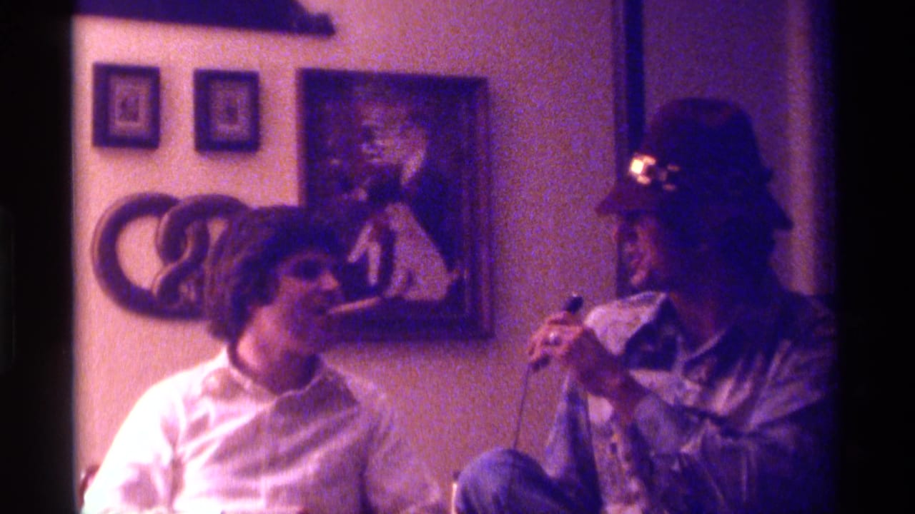 A seated man leans toward a cigar while another man in a hat holds a microphone.