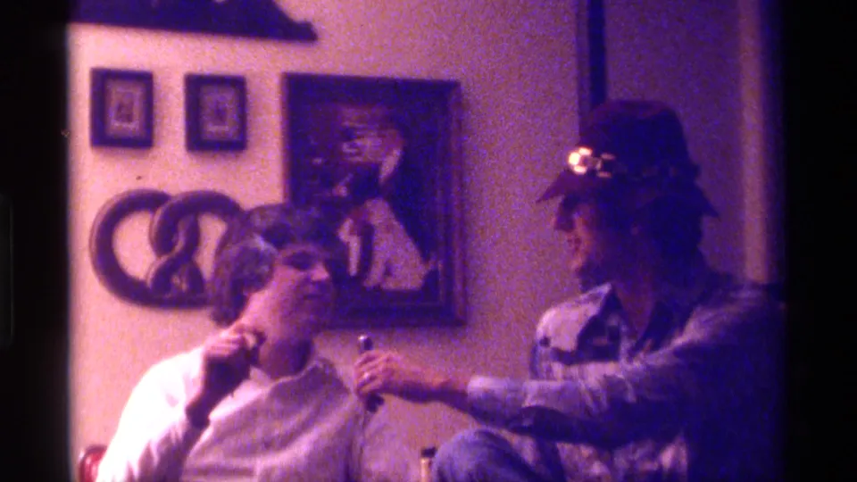 In the Kentucky home-movie scene, one adult man takes a drag from a cigar as his friend offers the microphone from beside him.