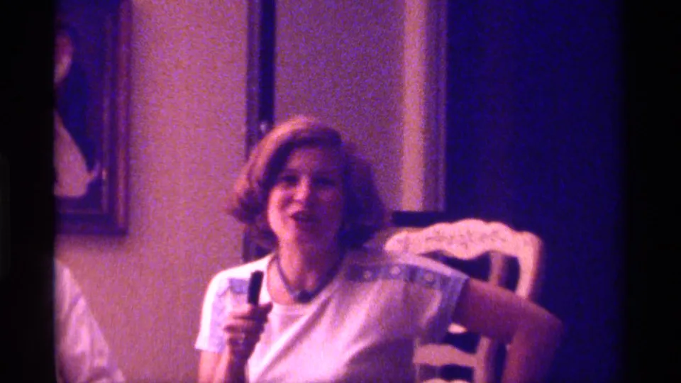 1976 Kentucky 8mm frame of a young woman with a short styled bob holding a wired microphone in a purple-blue lit interior.