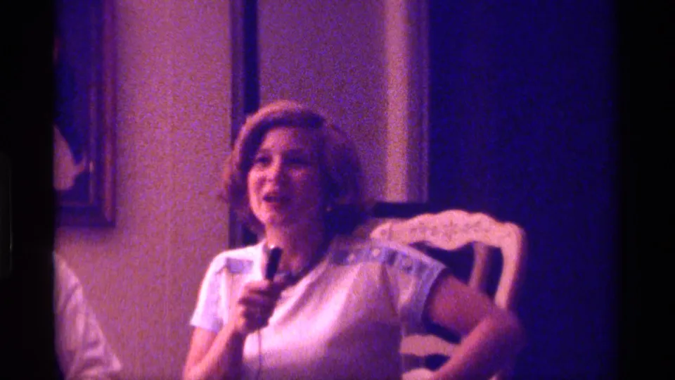 A smiling young woman in a white shirt with blue trim holds a microphone during an informal 1976 Kentucky home-movie performance.