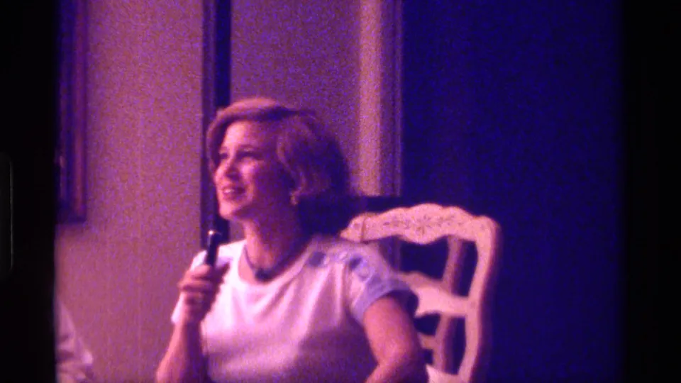 The young woman turns and smiles toward someone off camera, seated against an ornate white chair in a color-shifted 8mm interior.