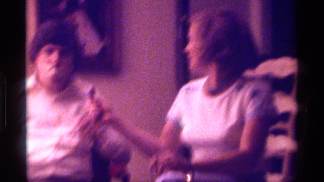 Two adults sit indoors, one with tape over his mouth, beside warm wall decor in soft purple 8mm color.