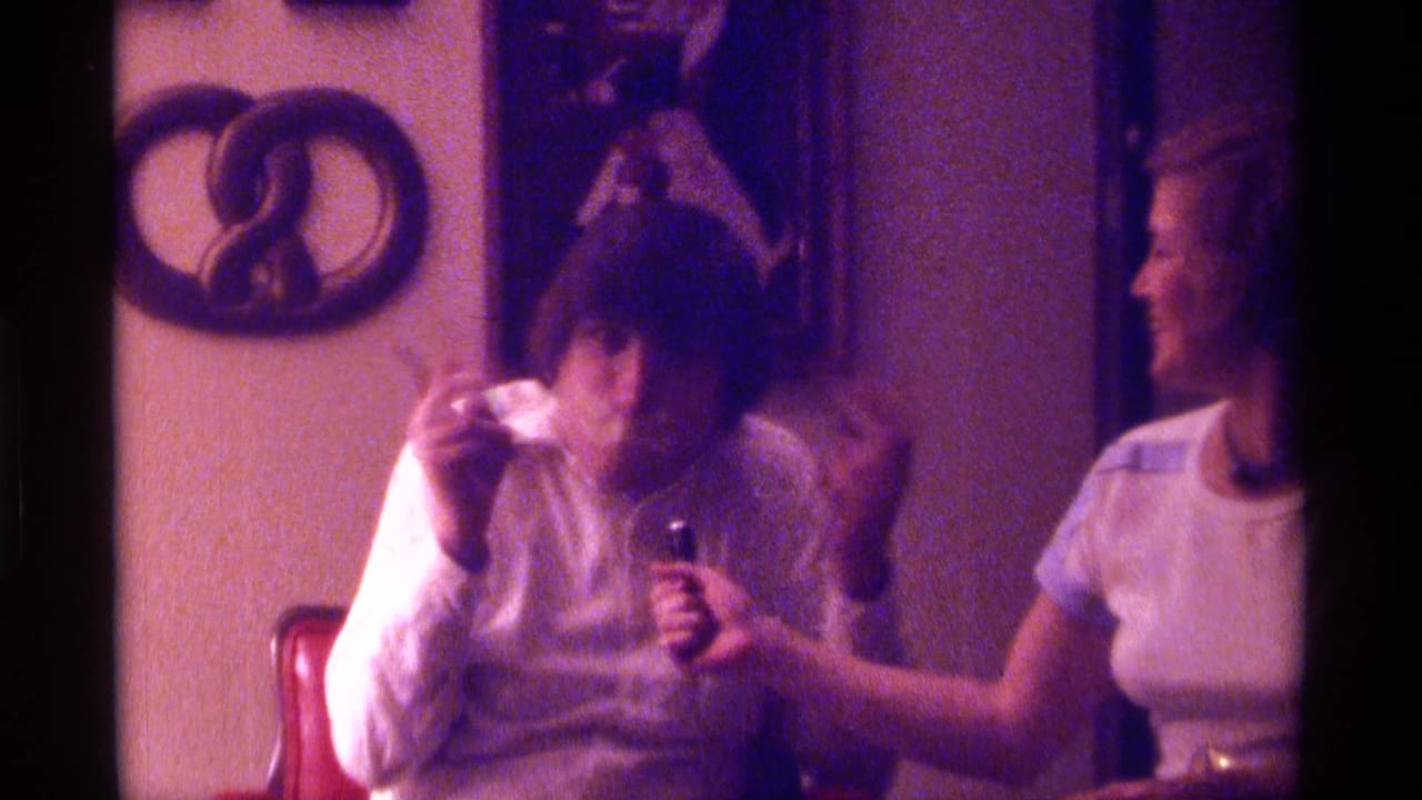 1976 Kentucky frame of a seated man with tape over his mouth while a woman holds a microphone toward him in a private interior.