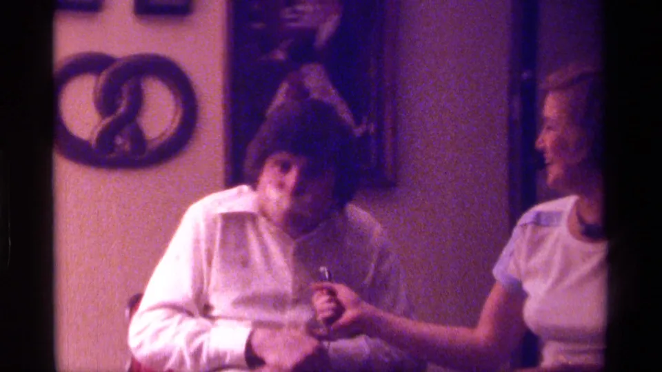 1976 Kentucky home-movie still of a man in a dark hat facing camera with tape over his mouth and a microphone nearby.
