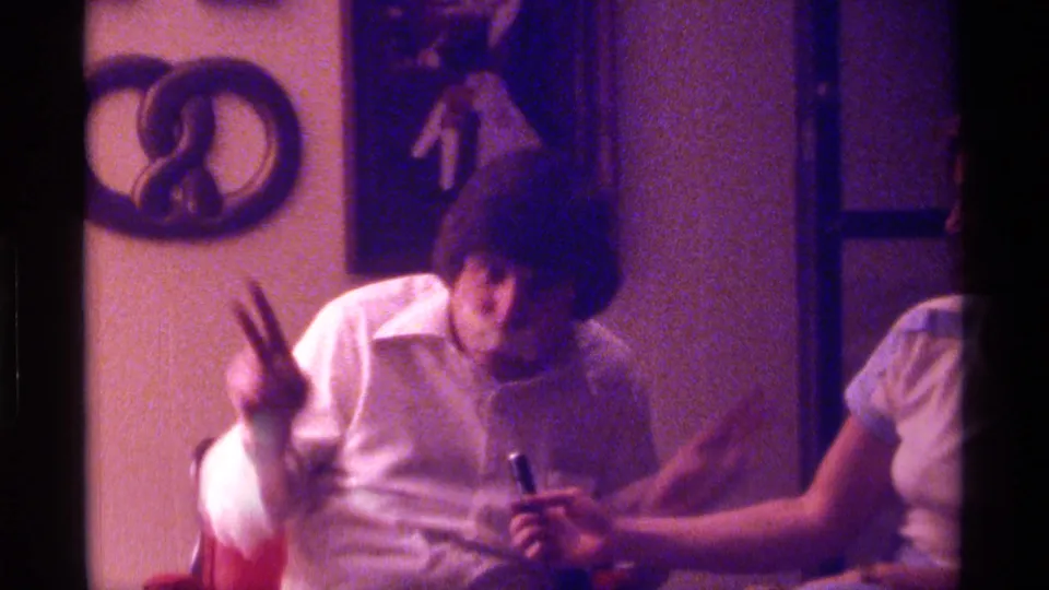 1976 Kentucky frame showing a taped-mouth man making a two-finger gesture as a woman holds a microphone in a dim private room.