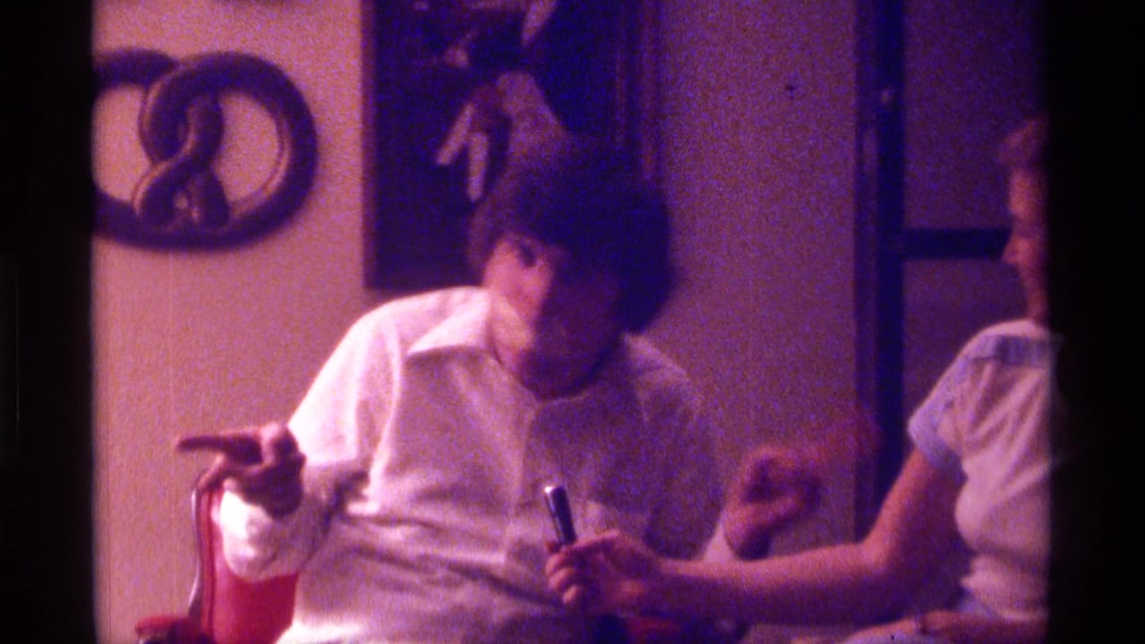 Young man in a white shirt gestures while seated at a table beside a woman holding a small object.