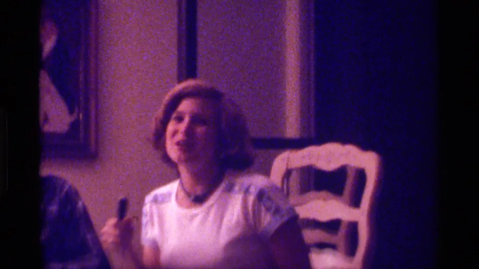 A woman in a light top sits in a private Kentucky home interior, holding a small microphone-like object during the playful 1976 scene.