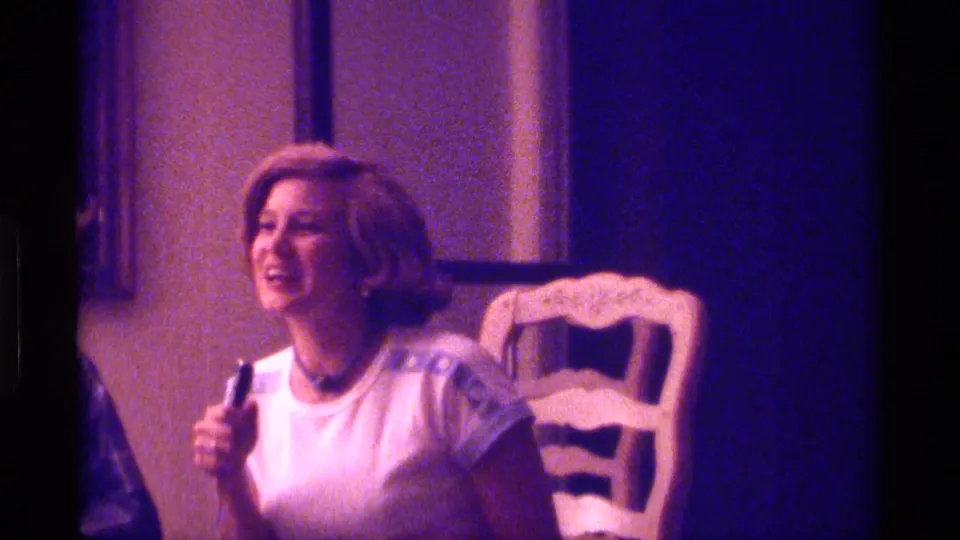 The woman laughs in the Kentucky 1976 home interior, holding a small microphone-like object beside a white dining chair.