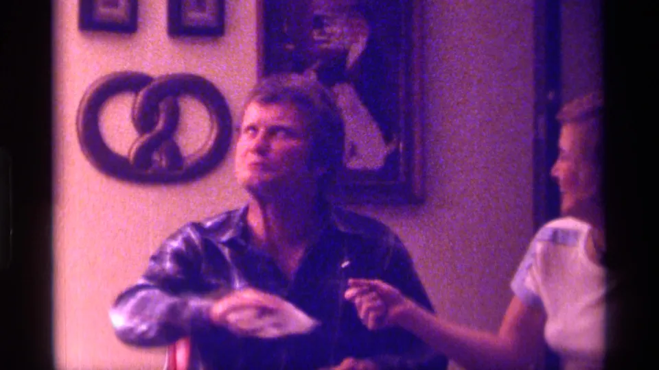 1976 Kentucky home-movie still of a man in plaid looking upward beside a woman, with wall art and warm domestic decor visible.