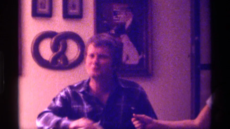 1976 Kentucky frame showing a seated man in plaid near a partially visible woman in a warm private interior with decorative wall art.