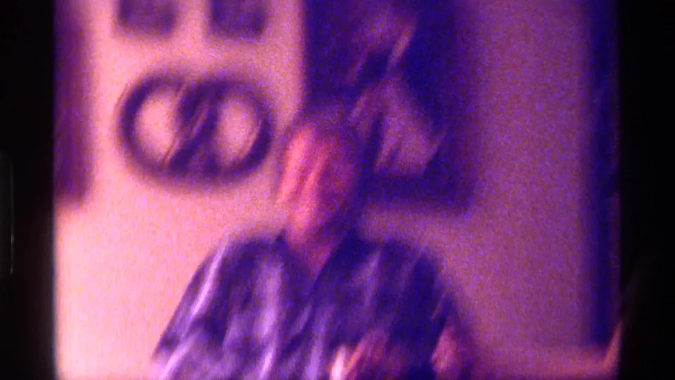 Blurred 1976 Kentucky 8mm frame showing an adult in plaid near wall decor inside a private home.