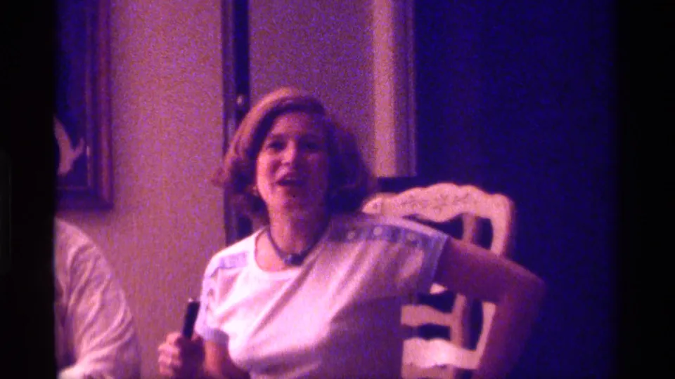 1976 Kentucky home-movie frame of a woman seated at a dining table area, smiling and holding a small dark object.