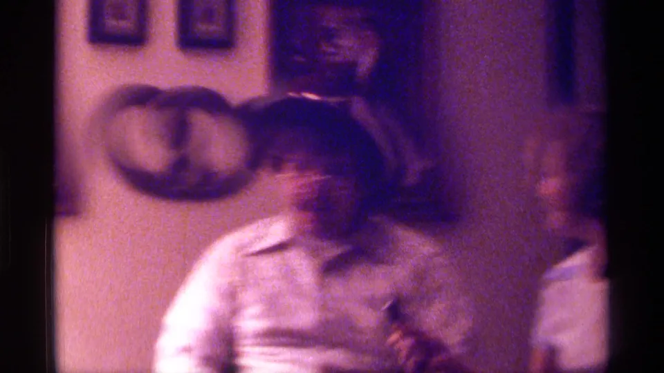Soft 1976 Kentucky home-movie frame of a seated man in plaid with wall decorations behind him.