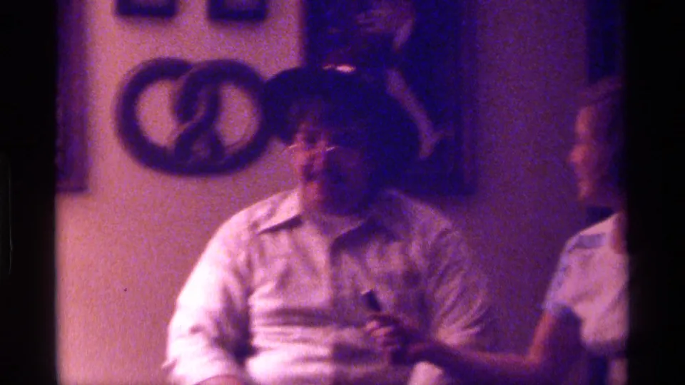 In a 1976 Kentucky home-movie frame, a man in a felt hat and light shirt sits in a dim interior with wall hangings behind him.