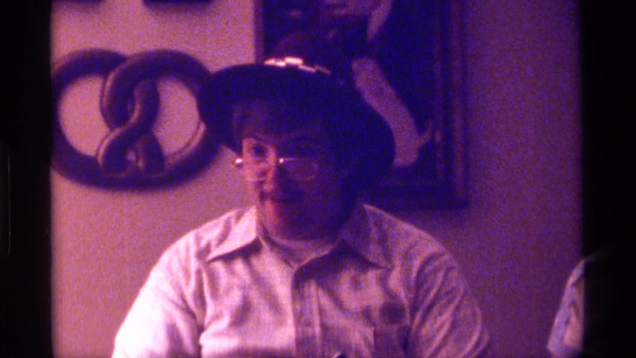 The man faces forward in the purple-tinted 8mm frame, with eyeglasses, a light shirt, and domestic wall decor visible behind him.