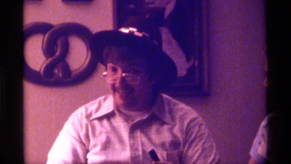 A 1970s Kentucky home-movie frame shows the man turned slightly to one side, seated in front of a wall hanging and framed decoration.