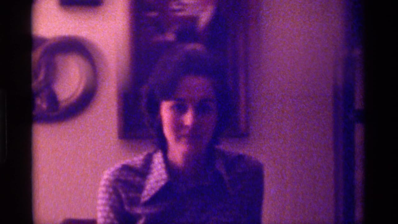 In 1976 Kentucky home-movie footage, a woman in a patterned blouse looks toward the camera against a wall with decor behind her.