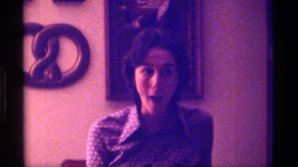 In 1976 Kentucky home-movie footage, a woman makes an animated open-mouthed face while seated or standing near indoor wall decor.