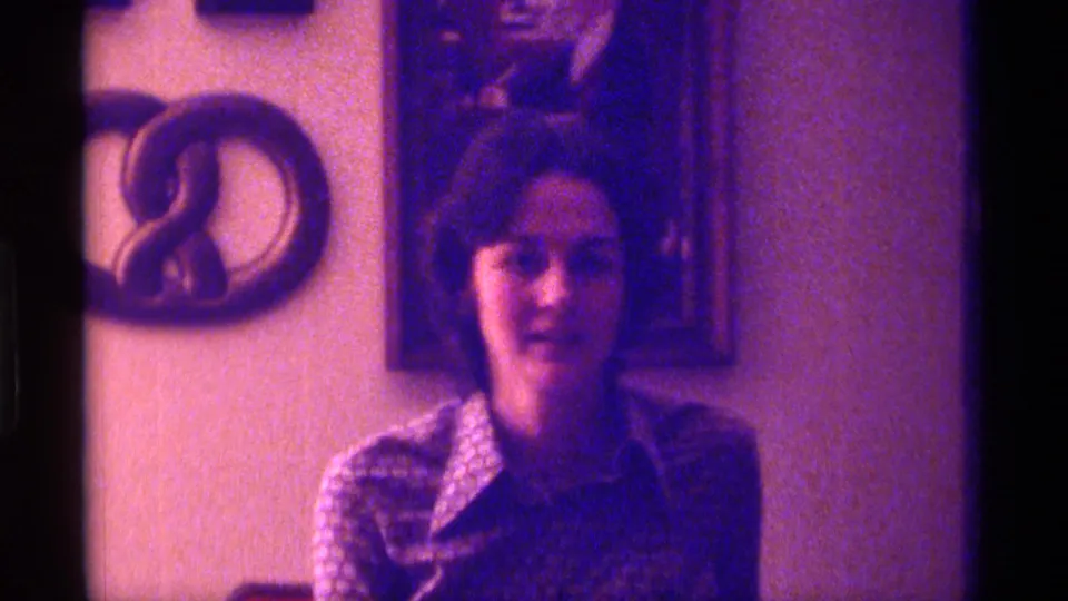In 1976 Kentucky home-movie footage, a woman in a patterned blouse looks toward the camera with a playful expression in a private interior.