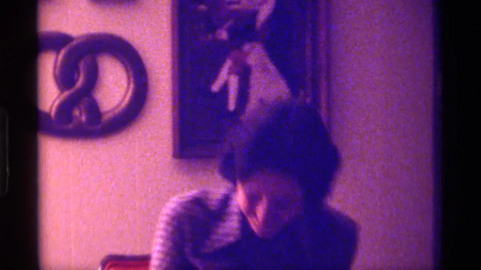 The woman leans forward during the candid Kentucky 1976 home-movie moment, with wall decor and a purple film cast visible.