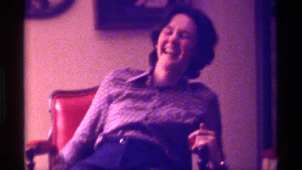 The seated woman laughs broadly in a red chair, captured in grainy 1976 Kentucky 8mm footage inside a private room.