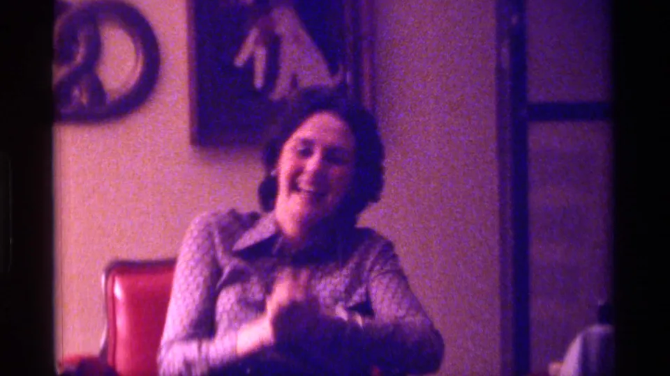 The woman continues laughing in the Kentucky 1976 interior, framed by a red chair, wall art, and soft magenta 8mm color.