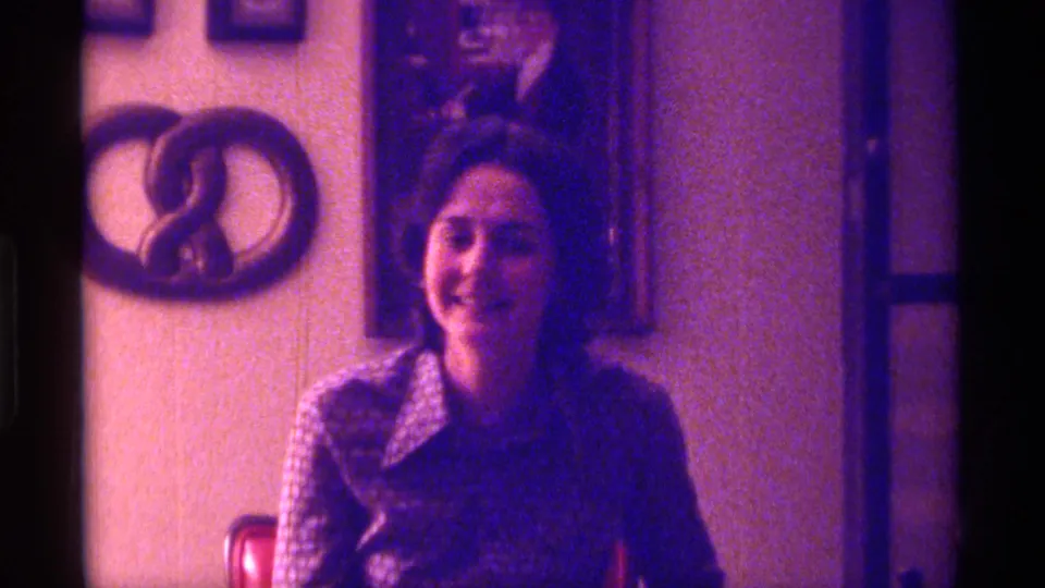 The woman remains seated and smiling in the Kentucky home setting, with framed pictures and a curved wall decoration visible behind her.