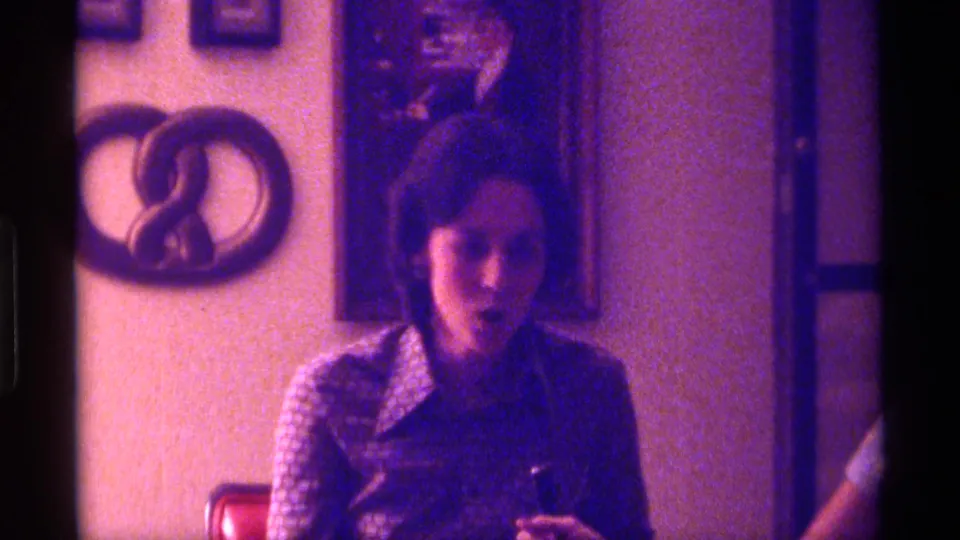 The woman appears to speak or gesture while leaning forward, framed by the red chair and wall decorations in the 1976 home-movie footage.