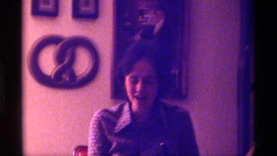 The seated woman looks downward in the Kentucky home interior, with a pretzel-shaped wall hanging and framed picture visible behind her.