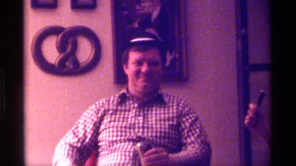 A man in a checkered shirt and cap sits in a Kentucky interior, holding a drink while using a microphone during a 1976 home movie moment.