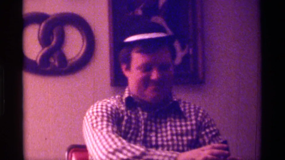 The man sits in the private Kentucky room, looking down with a drink in hand as the 8mm footage captures the informal exchange.