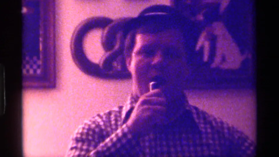 A close, soft-focus 1976 Kentucky home-movie frame shows the man raising a microphone near his mouth inside the decorated room.