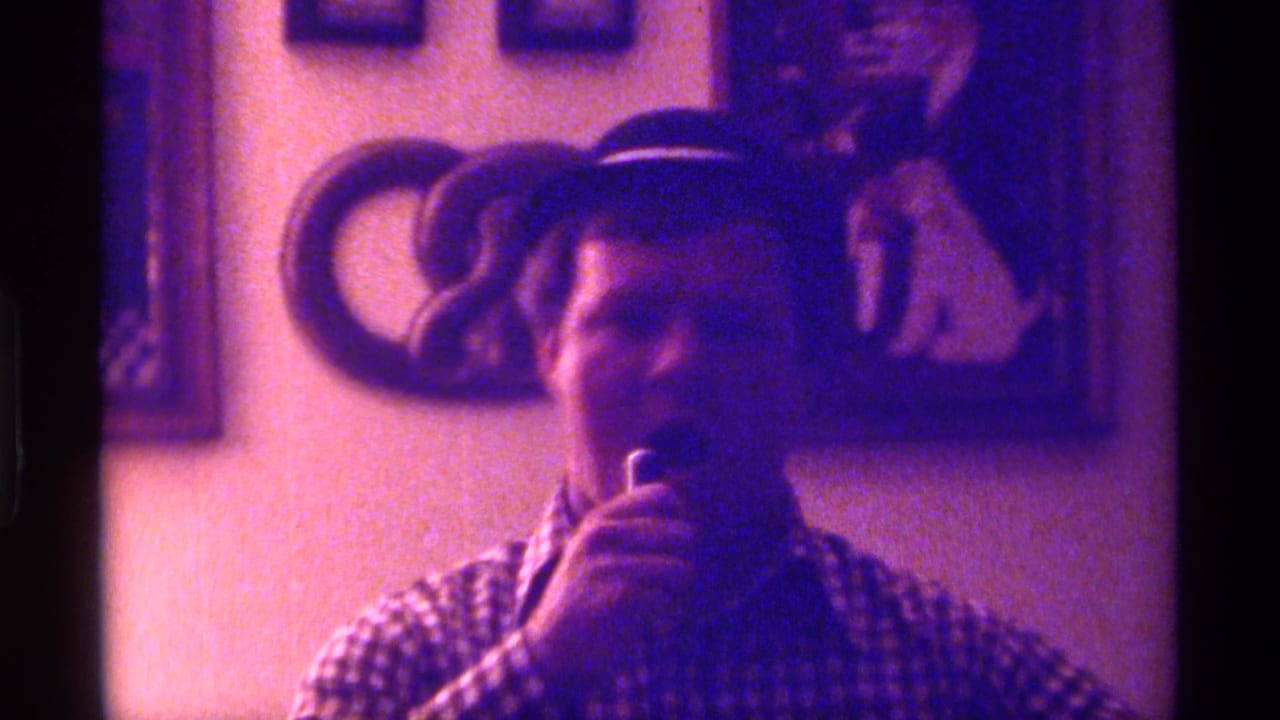 Man in a small dark hat holds a microphone indoors near framed wall art.