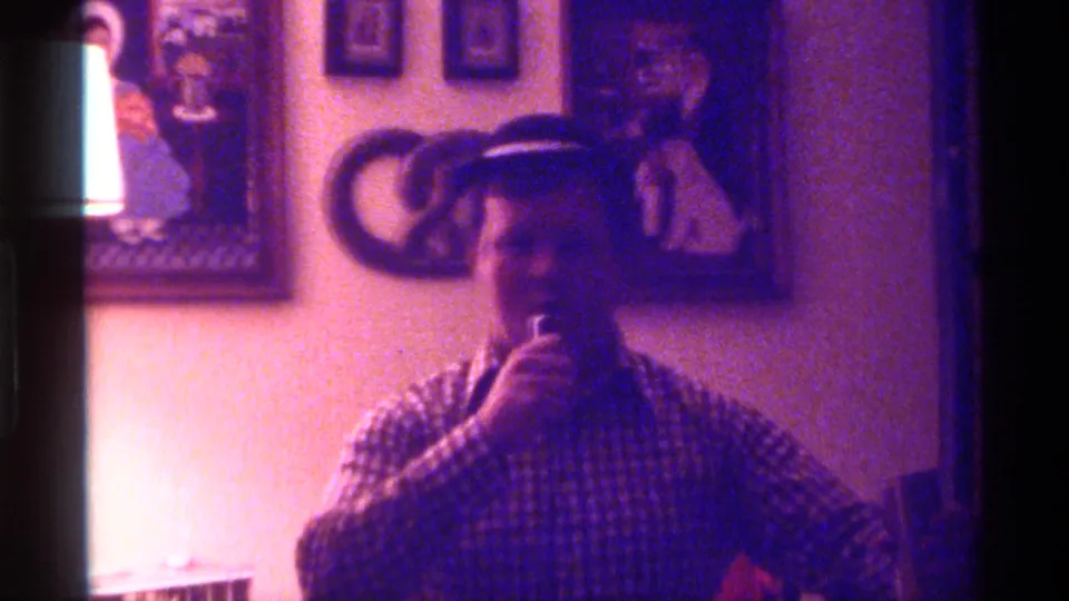A man in a dark hat and checkered shirt appears to speak or sing into a microphone in a Kentucky interior from 1976.