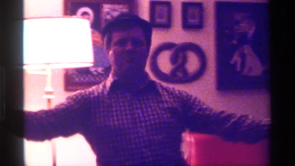 The man gestures with his arms while holding a microphone, with a bright lamp and framed wall art visible in the 1976 Kentucky home interior.