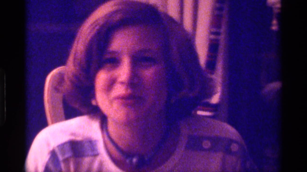 Young woman in a white shirt faces the camera in a dimly lit room.