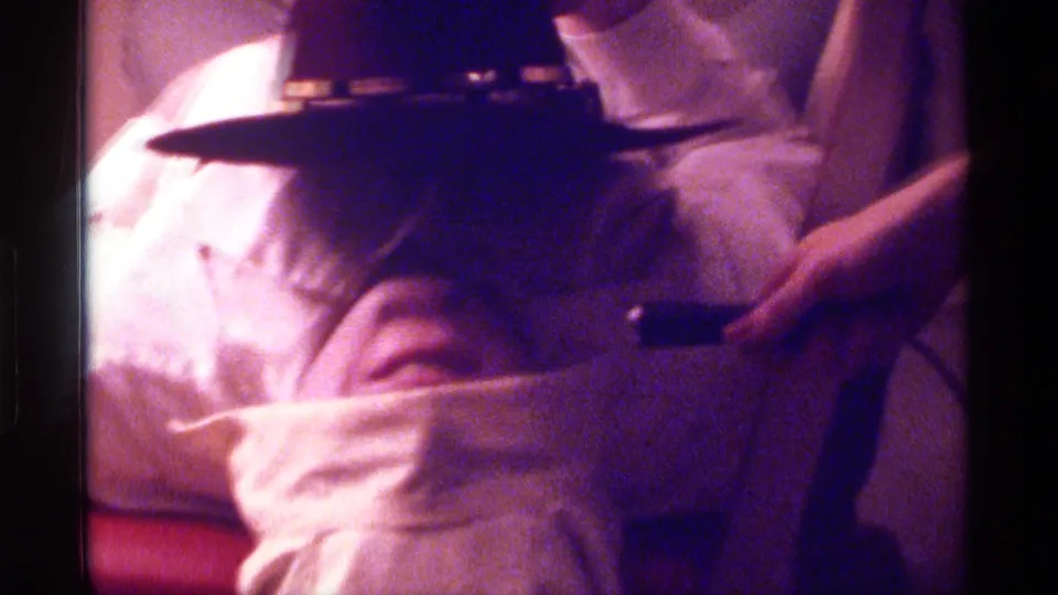 Reclining man appears blindfolded by a dark hat as another person's hand is visible beside him in the 1976 Kentucky home-movie scene.
