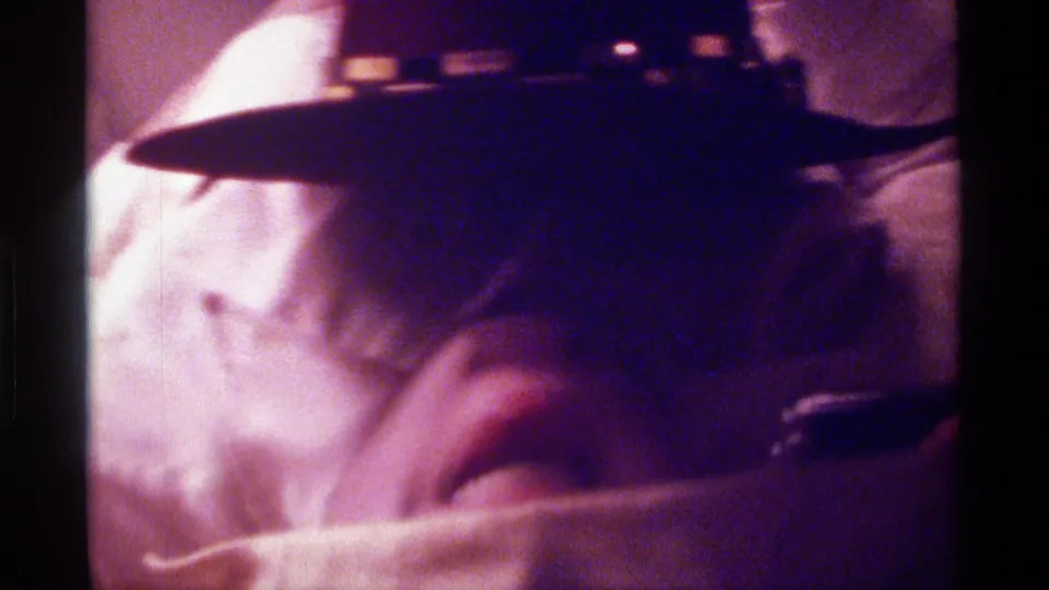 Kentucky 1976 home-movie frame of a person playfully covering their face with a white towel beneath a dark brimmed hat.