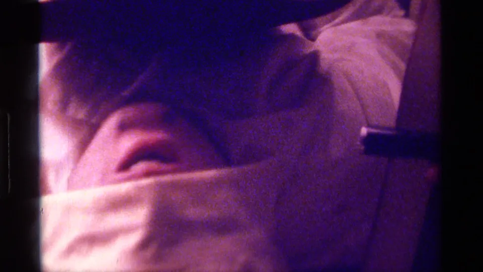 Purple-tinted 8mm close-up of the towel-and-hat gag, with the person's mouth visible as they face the camera.