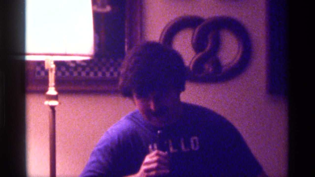 Man in a blue HELLO shirt holds a microphone in a dim room beside a bright floor lamp.