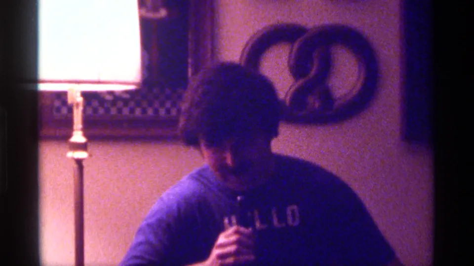 1976 Kentucky 8mm frame of a man holding a microphone in a dim private interior with a floor lamp and wall decor behind him.