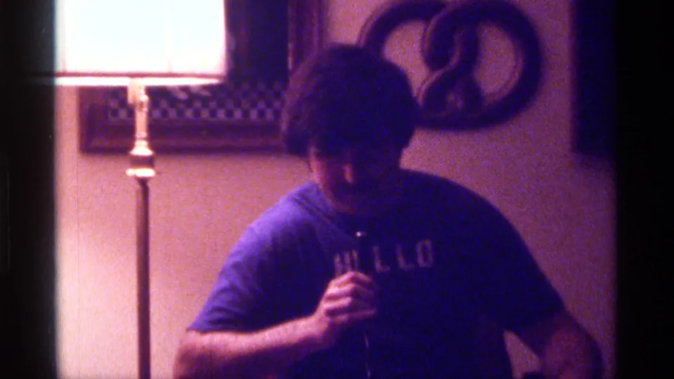 A dim 1976 Kentucky home-movie frame shows a man in a blue shirt holding a microphone with a lamp and wall hanging behind him.