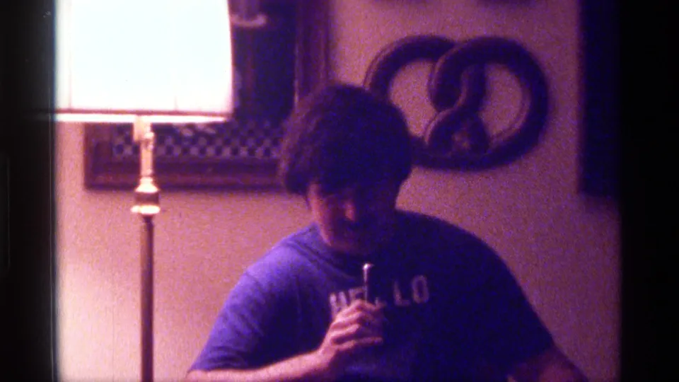 Purple-tinted 8mm frame from Kentucky in 1976 showing a man looking down with a microphone beside a bright floor lamp.
