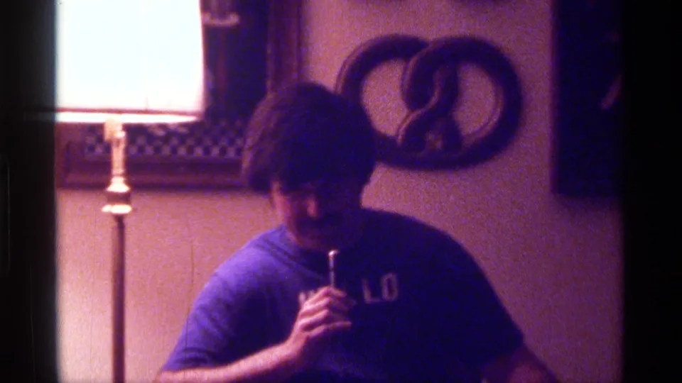 1976 Kentucky 8mm home-movie frame of a man with a microphone, blue HELLO shirt, floor lamp, and pretzel-shaped wall hanging.