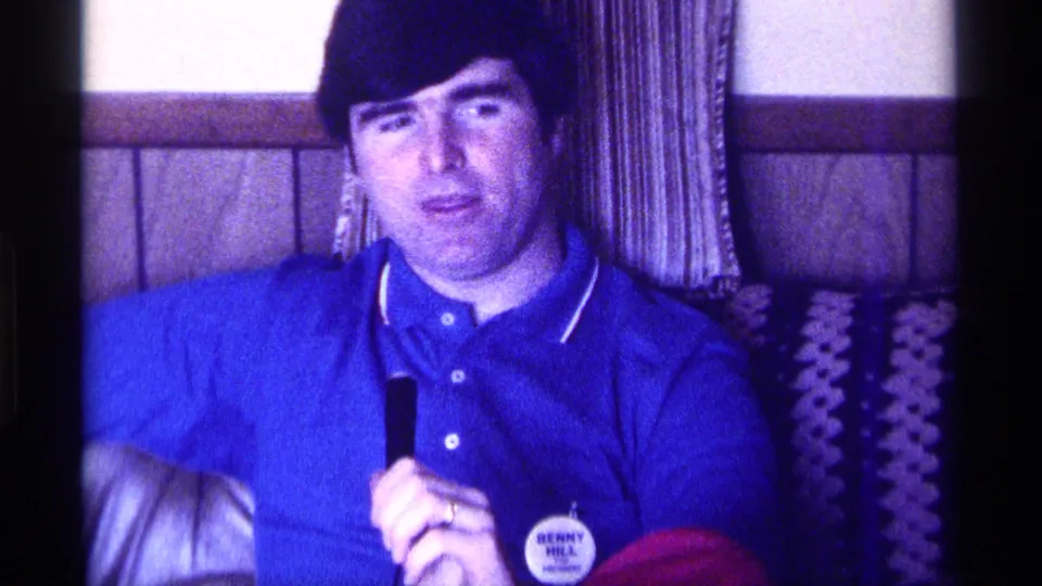 In 1976 Kentucky home-movie footage, a seated man in a blue polo shirt holds a microphone beside wood paneling and patterned textiles.