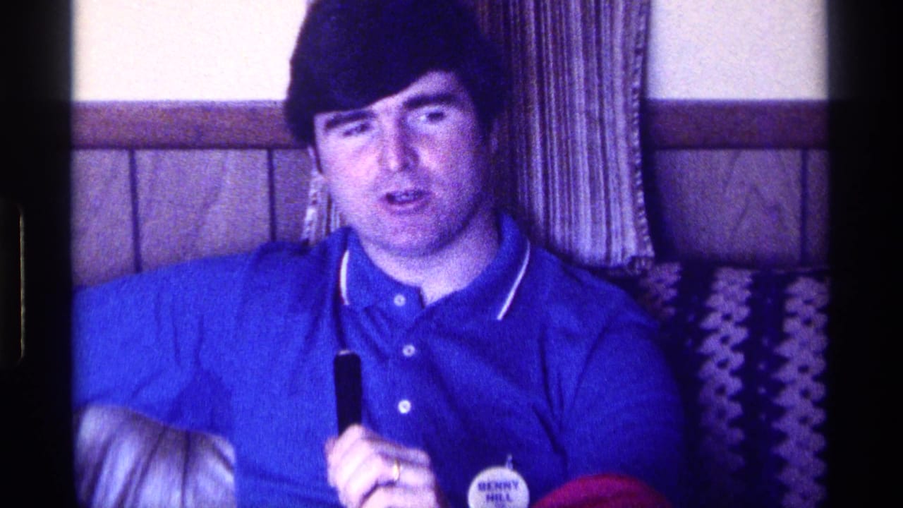 Man speaks into a handheld microphone while seated beside patterned upholstery.