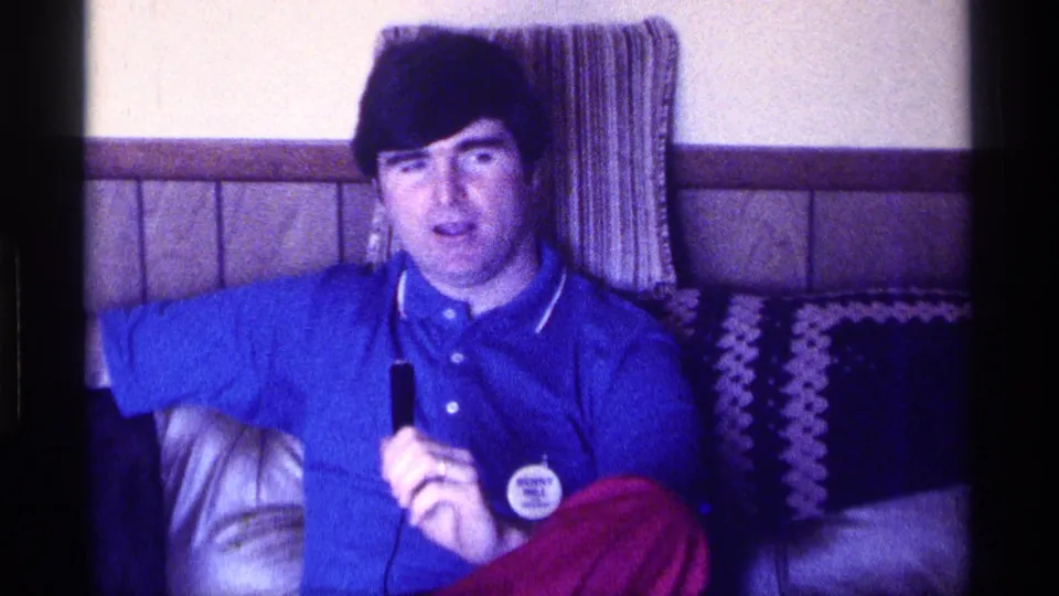 A wider frame shows the seated man with microphone, red pants, couch, and wood-paneled wall in a 1976 Kentucky living room.