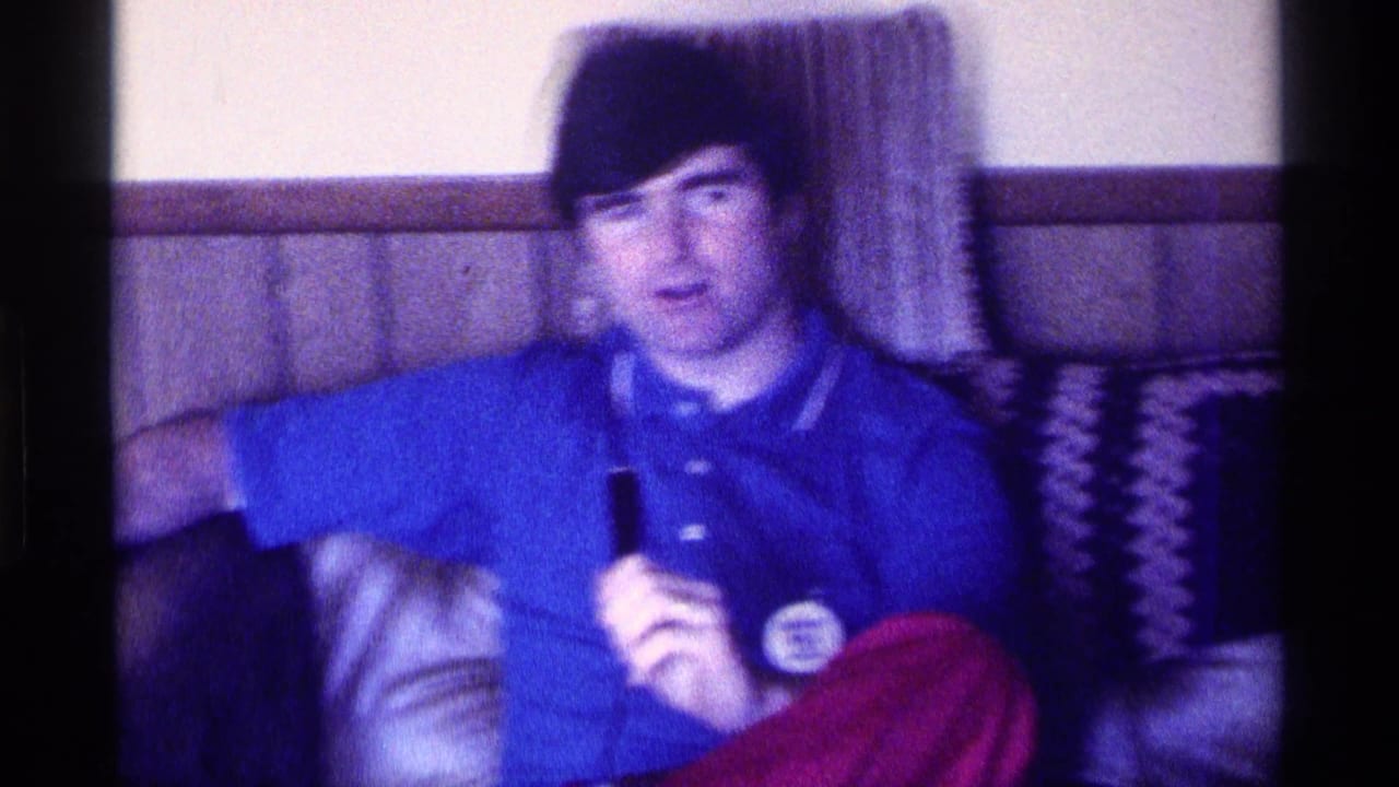 In a 1976 Kentucky home interior, a man in a blue polo and red pants sits on a couch holding a microphone.