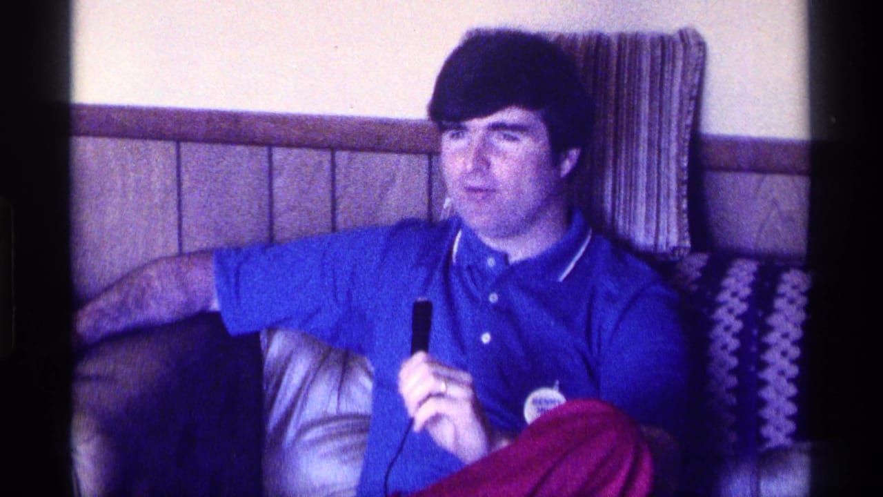 Man in a blue polo shirt sits on a couch with a microphone beside patterned cushions.